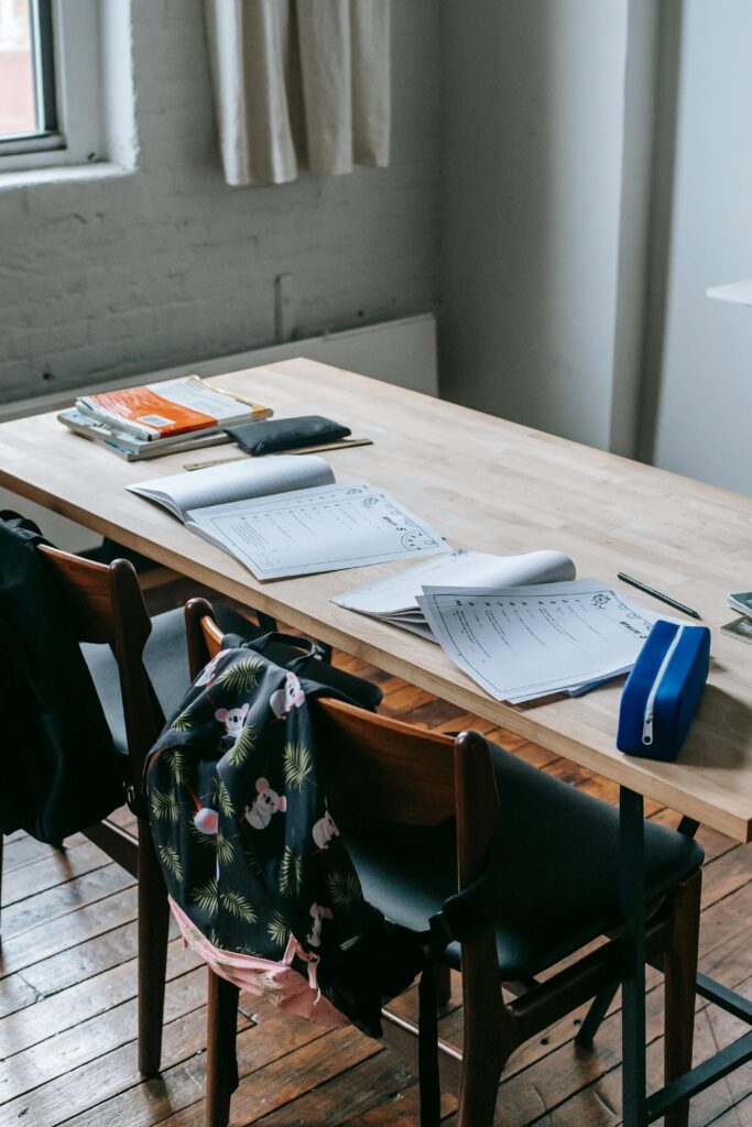 Wooden desk with opened copybooks and workbooks placed near chairs with backpacks in light classroom
